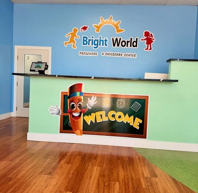 Reception area of Bright World Preschool and Childcare Center with a colorful sign, a cartoon pencil character with a welcoming gesture, and a chalkboard sign reading 'Welcome'.