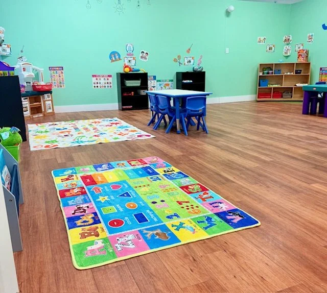 Empty children's playroom with colorful animal alphabet rug, small blue table with chairs, and shelves with toys and books.