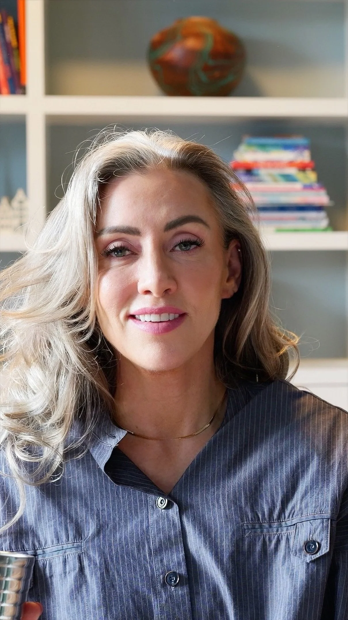 A woman with blonde hair and blue eyes smiling, sitting indoors with a bookshelf behind her containing colorful books and decorative items.