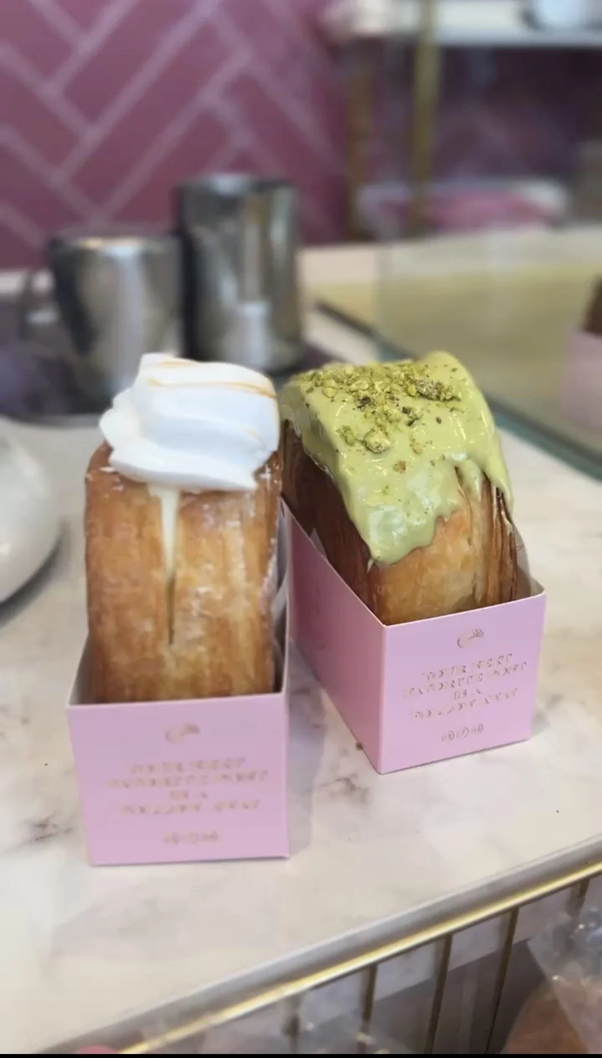 Two large pastry croissants filled with cream, one topped with white icing and whipped cream, the other topped with green glaze and crushed pistachios, placed in pink paper holders on a marble counter.