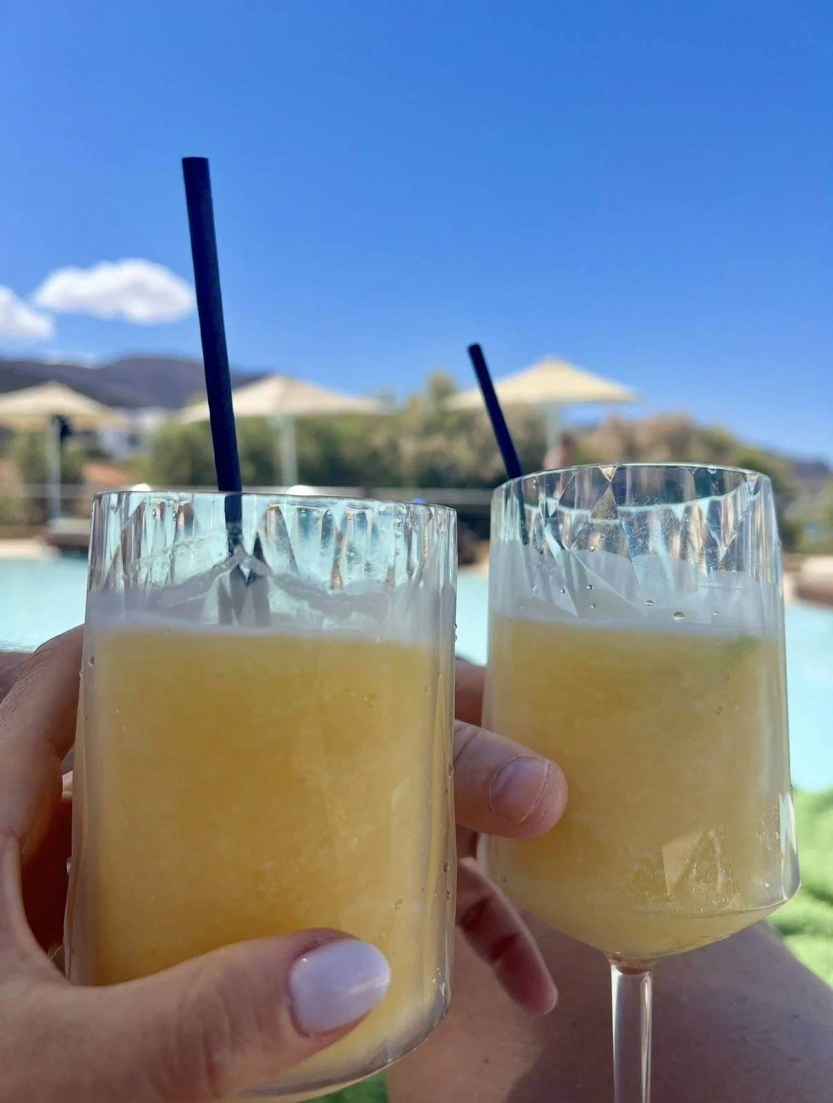 Two glasses filled with yellow-colored beverages, each with a straw, held up against a poolside background with blue sky, white clouds, and umbrellas.