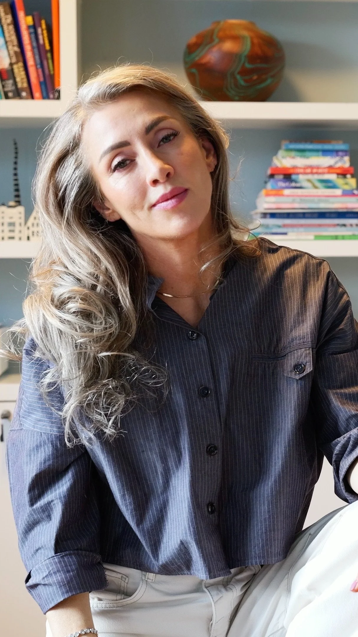 A woman with long, wavy hair, wearing a dark button-up shirt and light-colored pants, sitting in front of books and decorative items on a bookshelf.