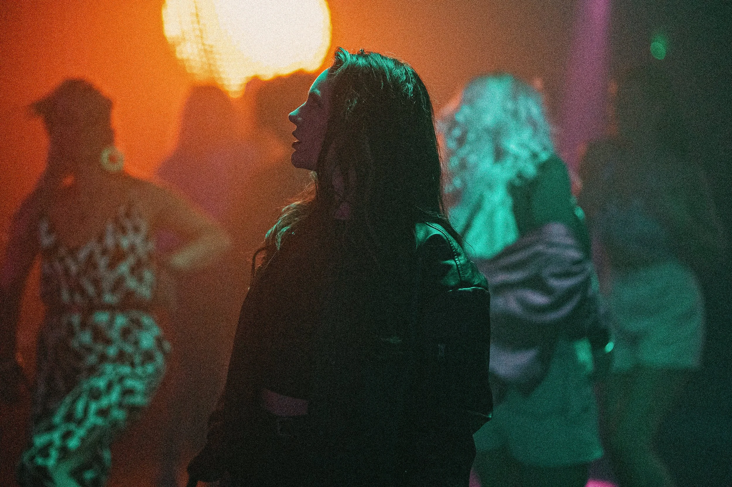 A woman with long dark hair in profile, dancing in a dimly lit club with colorful lights and other people in the background.