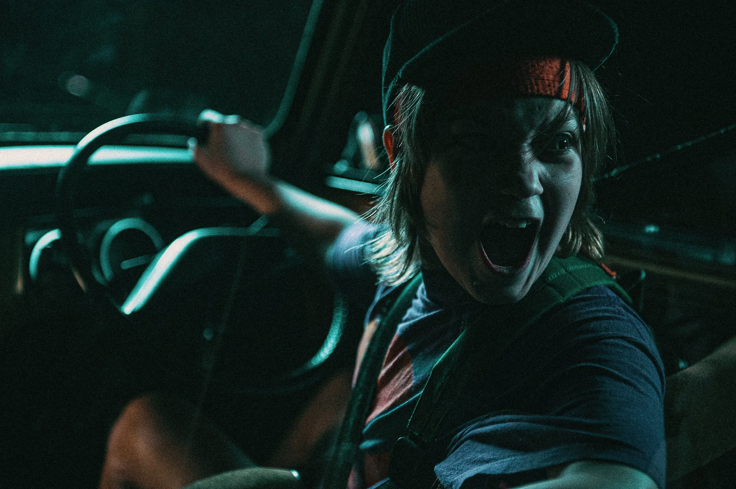 A woman with short blonde hair, wearing a dark cap and headband, appears to be shouting or yelling while holding a steering wheel in a dark, greenish-lit environment.