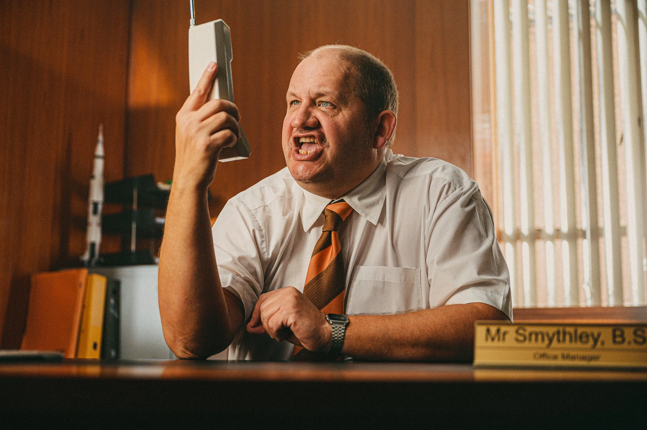 A middle-aged man dressed in a white shirt and orange striped tie sits at a desk in an office, angrily yelling and holding a telephone receiver near his mouth. The desk has a nameplate that reads 'Mr. Smythlow, B.S.', and the background features wooden walls and vertical blinds covering a window.