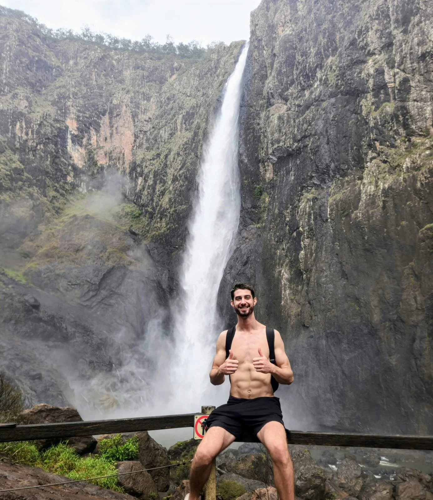 A smiling man in black shorts and a backpack sitting on a wooden fence in front of a waterfall, giving two thumbs up.