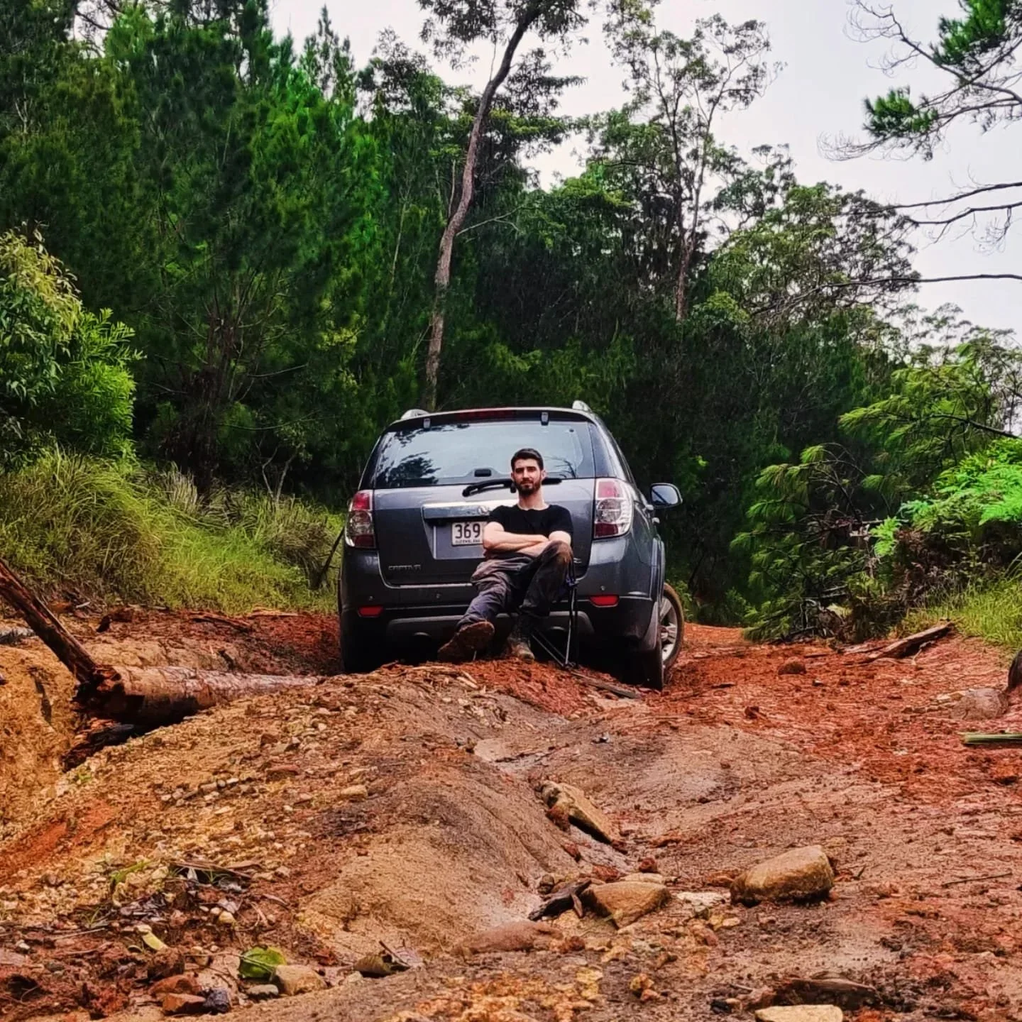 A man sitting in front of a black SUV on a rugged, dirt trail surrounded by green trees and foliage.