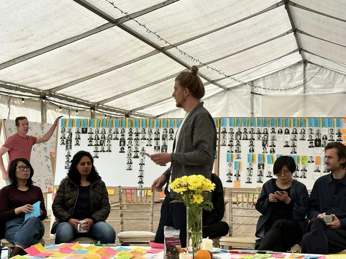 A group of people inside a large tent, some sitting and some standing, with a man in a gray blazer holding a paper. Behind him is a wall decorated with black-and-white portrait photographs and colorful sticky notes. There is a table with yellow flowe