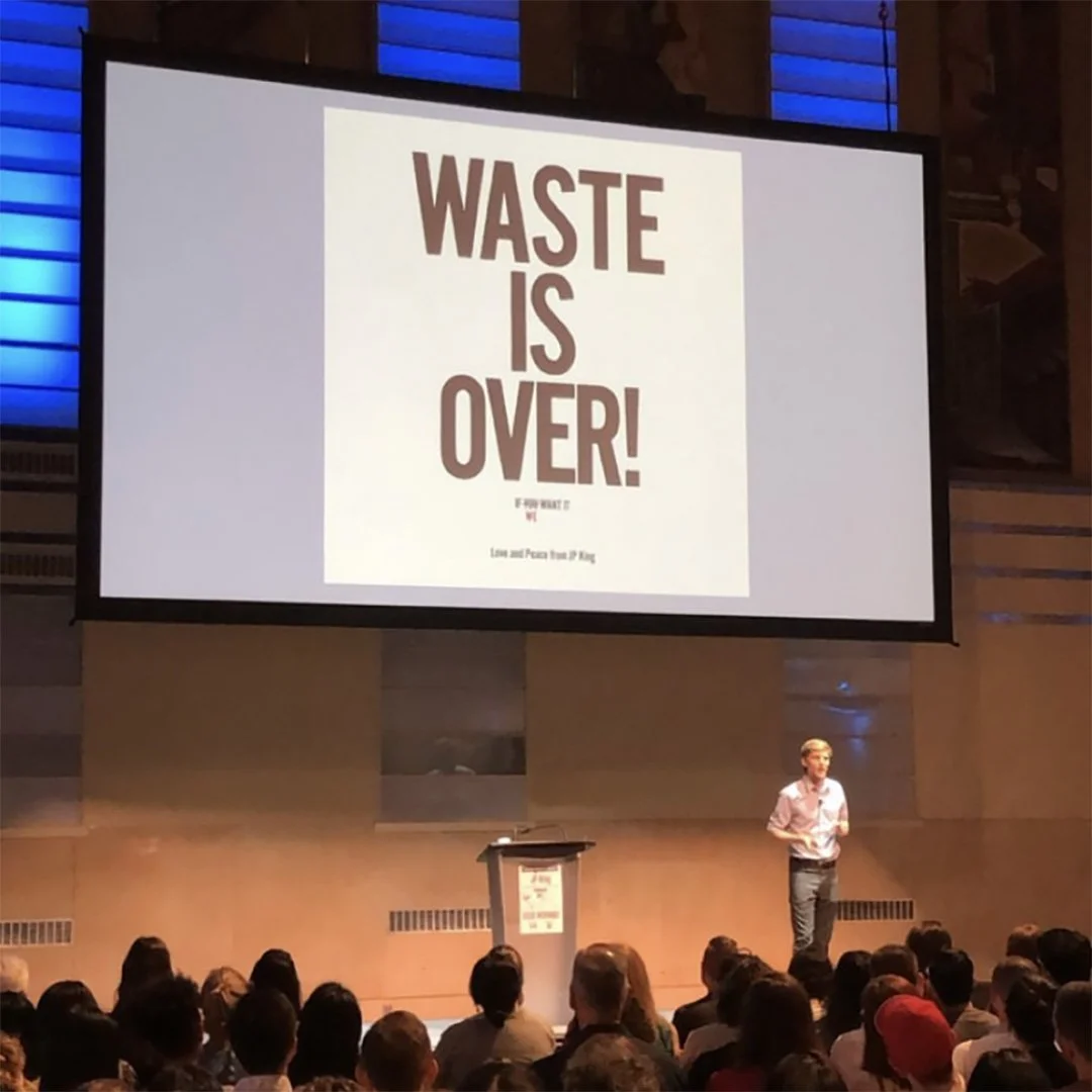 A presentation slide with the text 'Waste is Over!' in large, bold letters, displayed on a screen in front of an audience in a conference room.