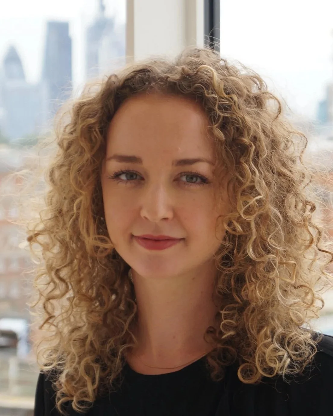 Close-up portrait of a young woman with curly blonde hair and light blue eyes, smiling softly in front of a large window with a cityscape view.