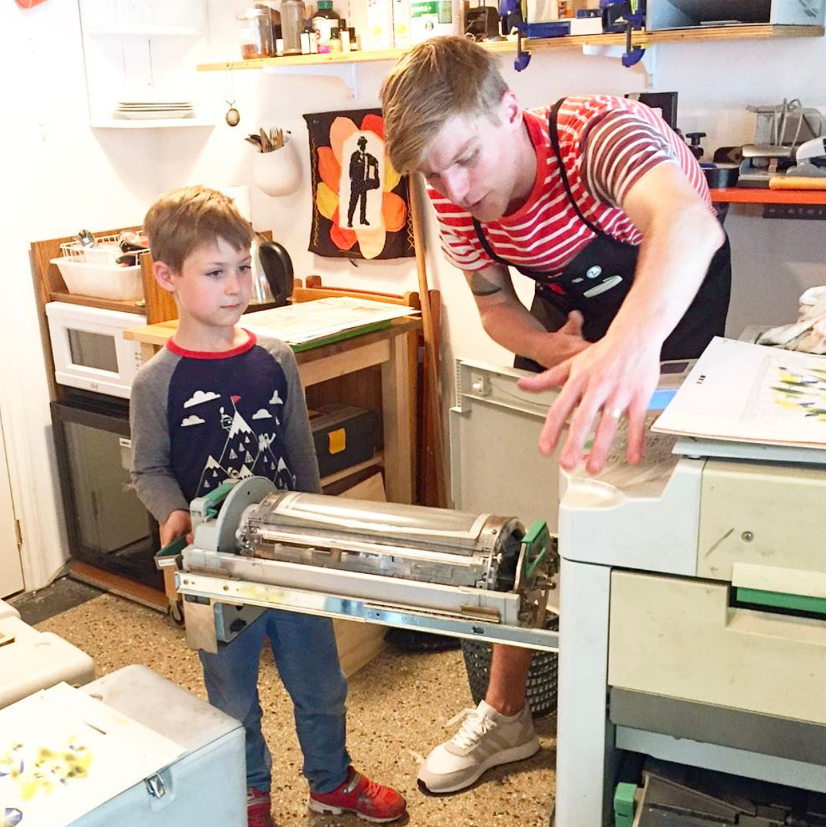 A man and a boy in a workshop. The boy is holding a machine part, and the man is explaining something, pointing at the machine on the workbench.