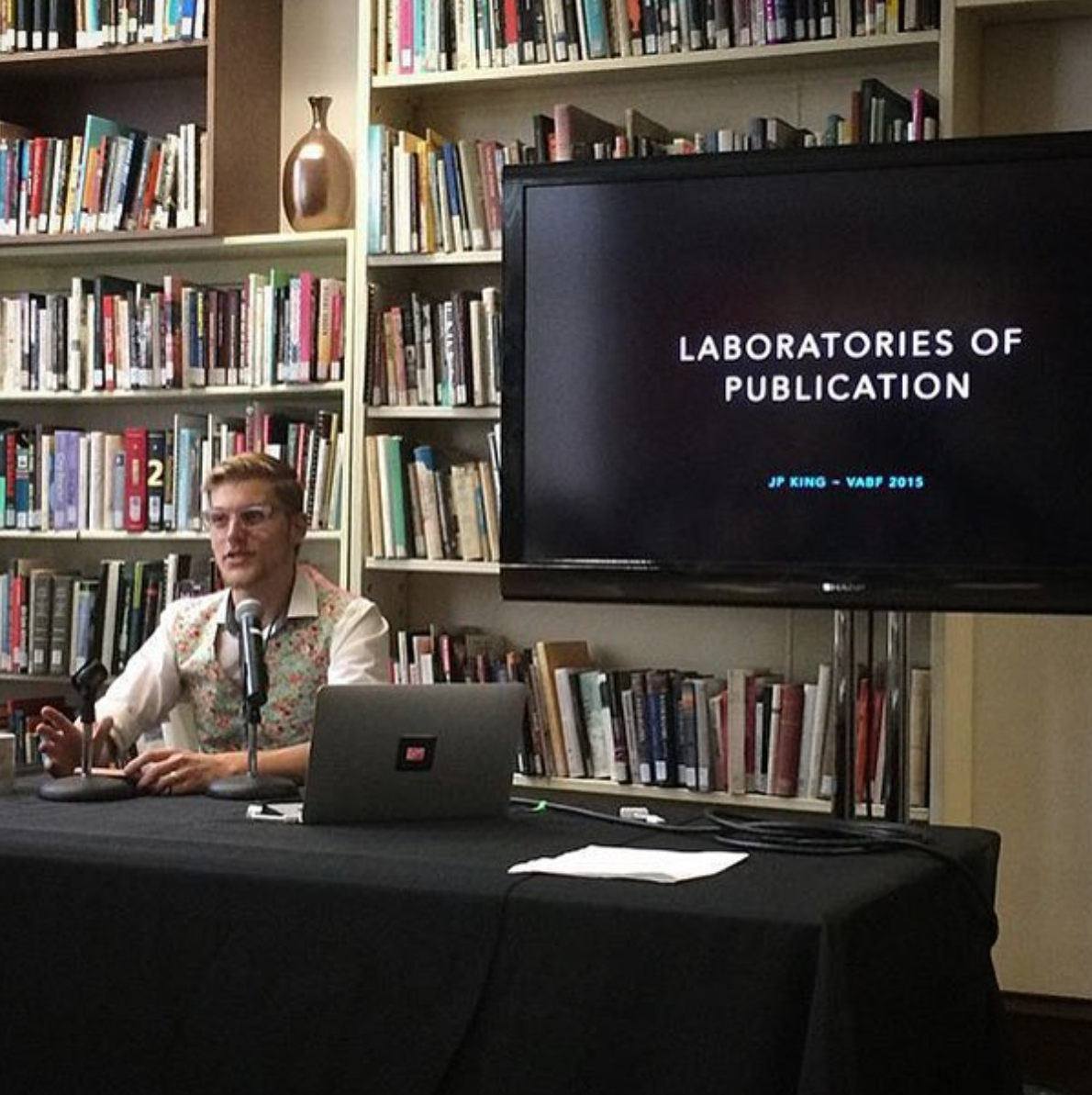 A person sitting at a table with microphones, a laptop, and papers, speaking in front of a large screen displaying 'Laboratories of Publication' and 'JP King - VABF 2015,' with bookshelves filled with books in the background.