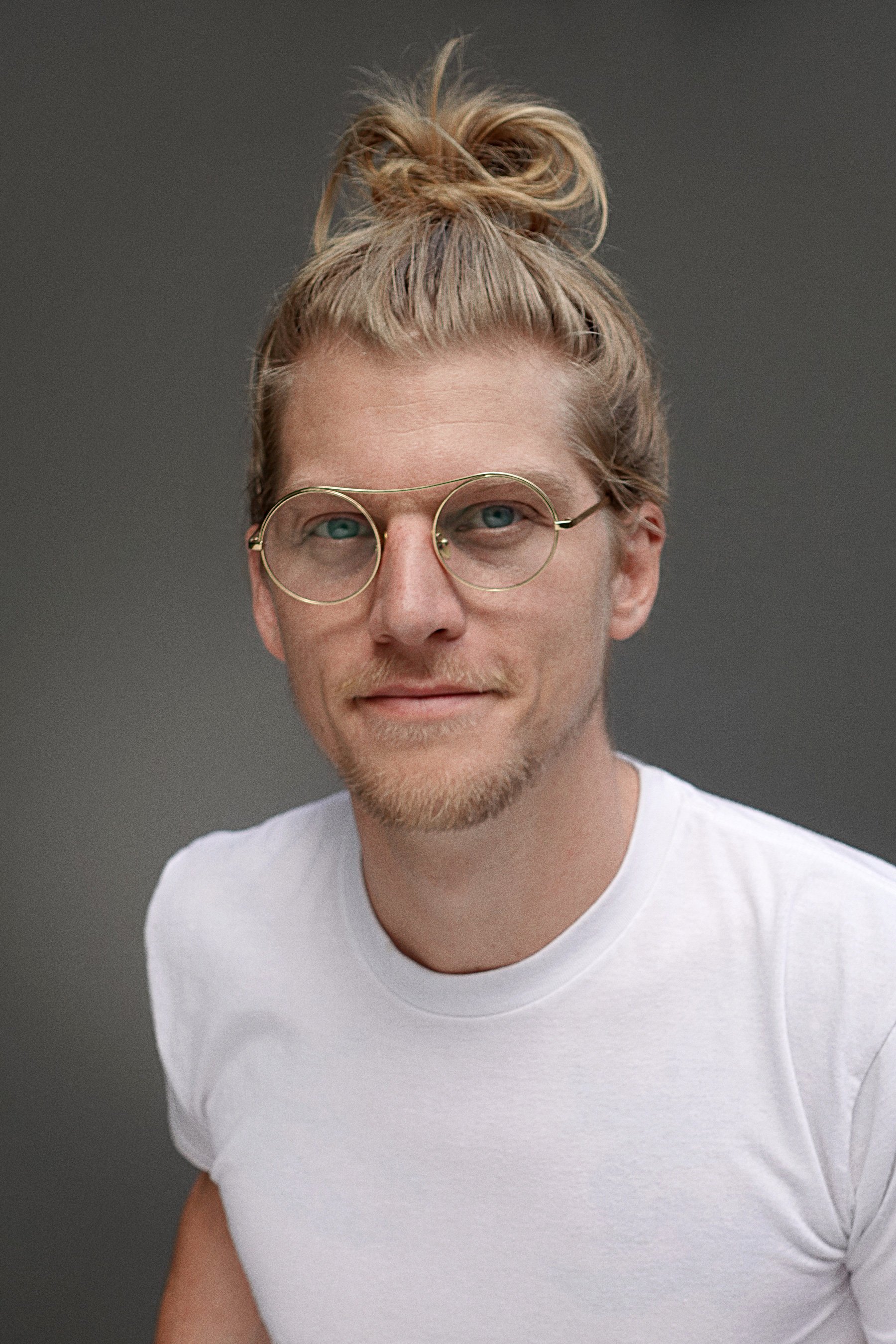 A young man with light brown hair tied in a messy bun, wearing round glasses and a white t-shirt, looking at the camera with a neutral expression against a gray background.