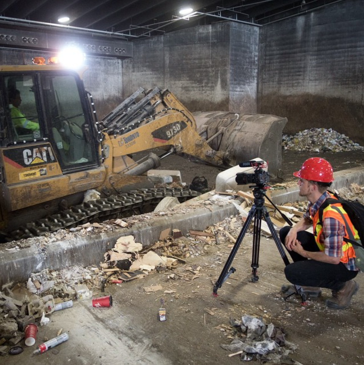A construction worker with a red safety helmet and safety vest is crouching next to a tripod-mounted camera inside a tunnel, filming or photographing construction equipment that is digging into the ground. The scene shows debris, trash, and construct