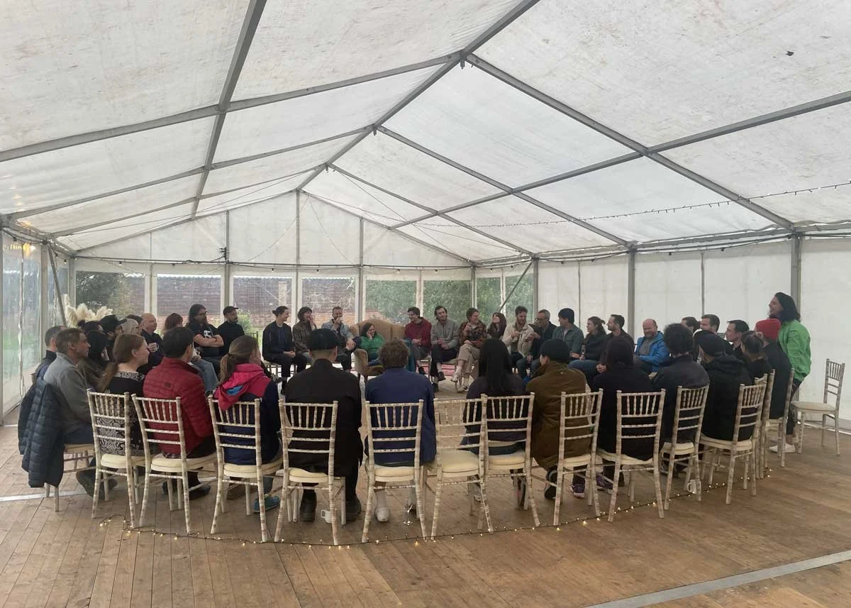 A large group of people sitting in a circle inside a white tent, participating in a discussion or meeting.