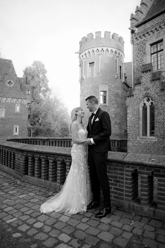 Ein Brautpaar in formeller Kleidung vor einer alten Burgmauer, vermutlich bei einer Hochzeit, in schwarz-weiß aufgenommen. Die Hochzeit findet im Schloss Paffendorf in Bergheim, Nähe Köln statt. 