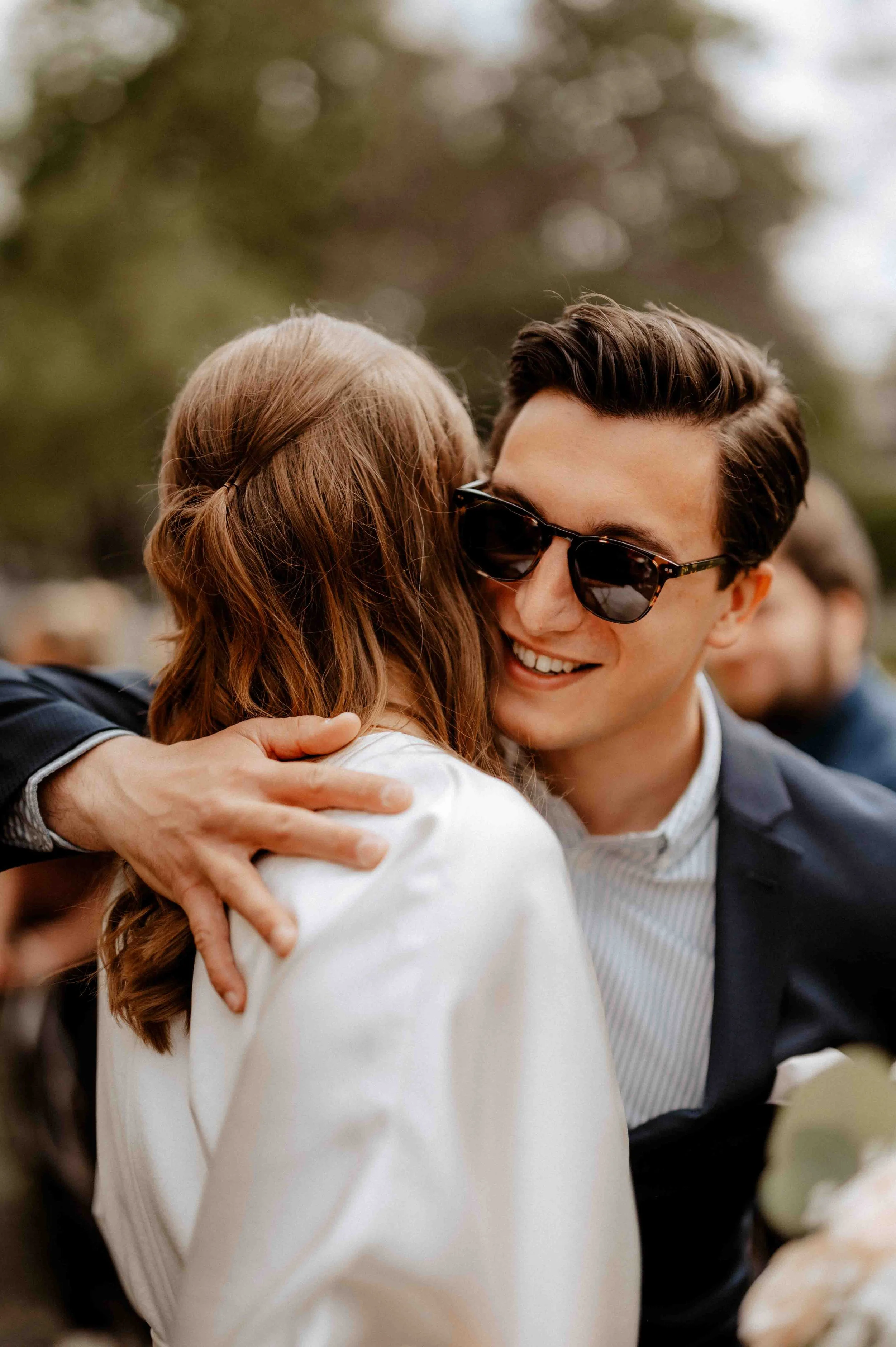 Ein Mann trägt dunkle Sonnenbrille und umarmt eine Frau. Beide lächeln. Der Hintergrund ist unscharf mit Bäumen, wahrscheinlich im Freien bei einer Feier. Die Hochzeit findet in Düsseldorf, Nähe Köln statt. 