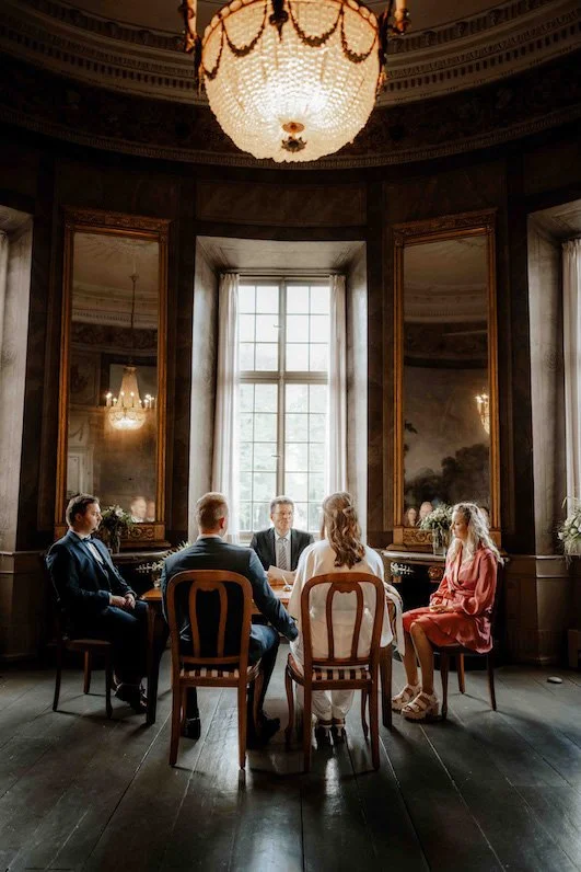 Sechs Menschen sitzen um einen runden Tisch in einem eleganten, historischen Raum mit hohen Fenstern und großen Spiegeln, beleuchtet von einem prunkvollen Kronleuchter im Haus Cromford in Ratingen, in der Nähe von Düsseldorf. Es handelt sich um die T