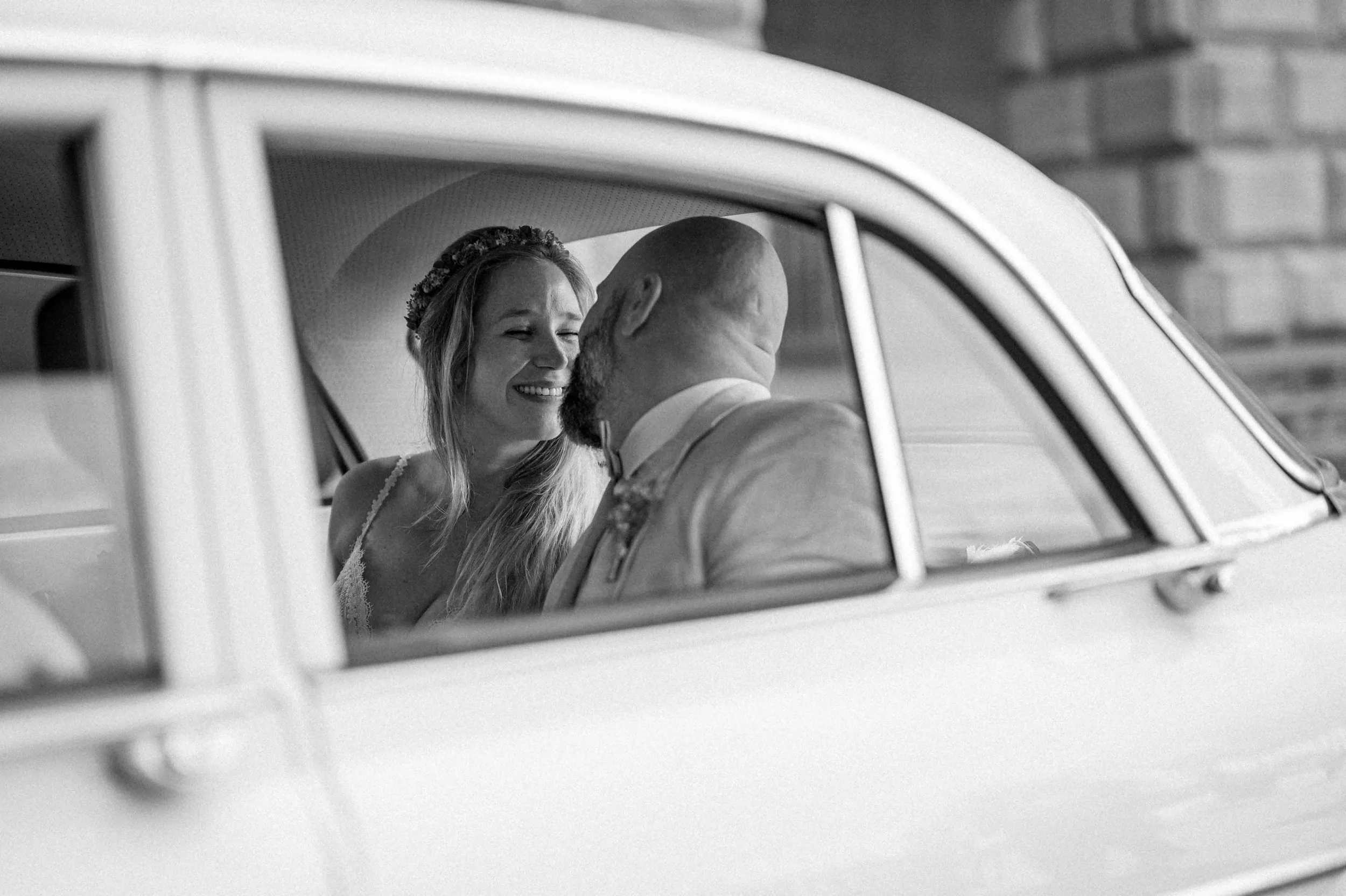 Schwarz-weiß-Foto eines Paares in einem Auto, das an ihrem Hochzeitstag fährt, die Braut lächelt glücklich. Die Hochzeit findet in Mülheim an der Ruhr bei Düsseldorf statt. 