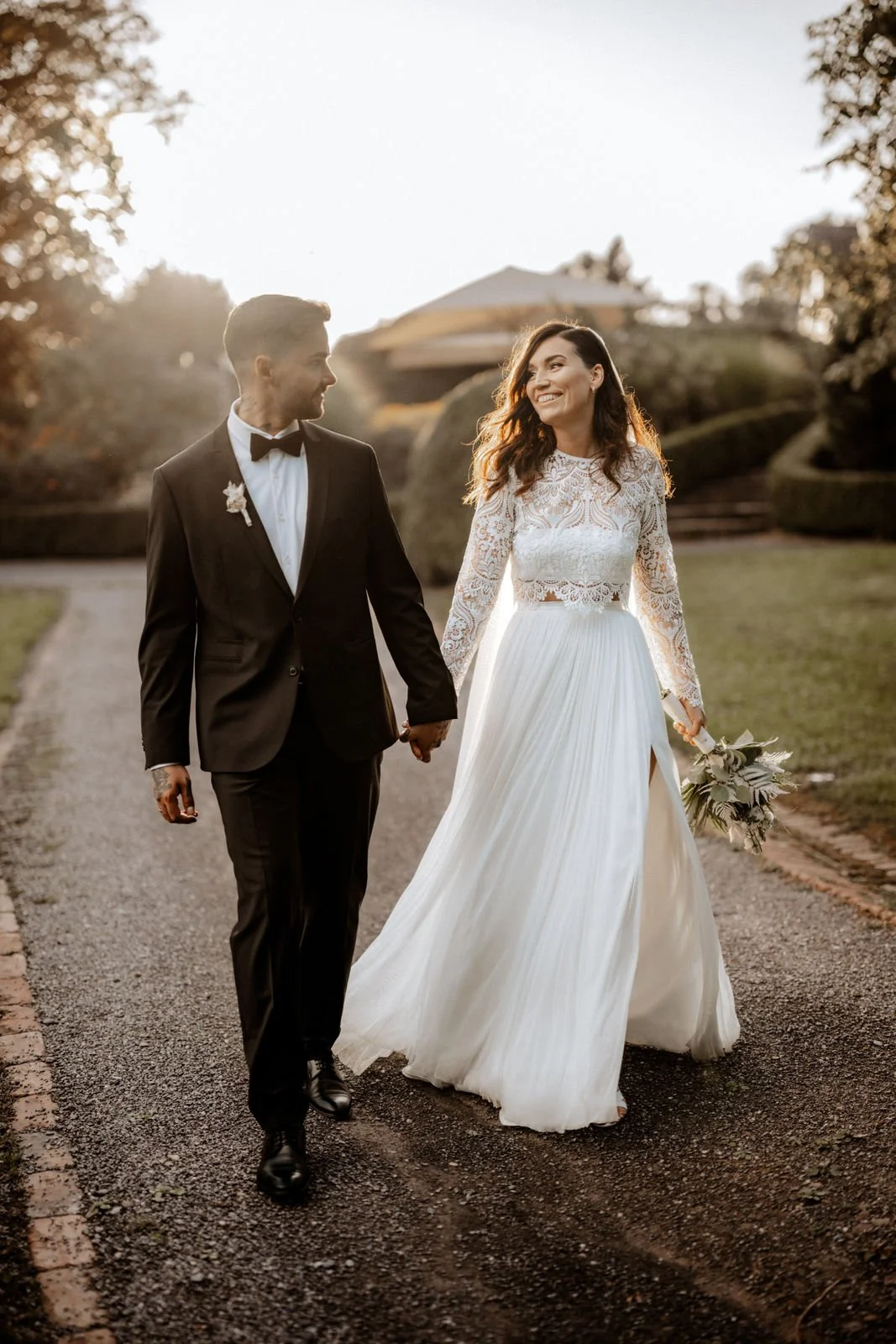 Ein Brautpaar in formeller Kleidung, die Hand in Hand auf einem Weg in einem Garten bei Sonnenuntergang spazieren geht. Die Hochzeit findet in Solingen im Schloss Grünewald, Nähe Köln und Düsseldorf statt. 