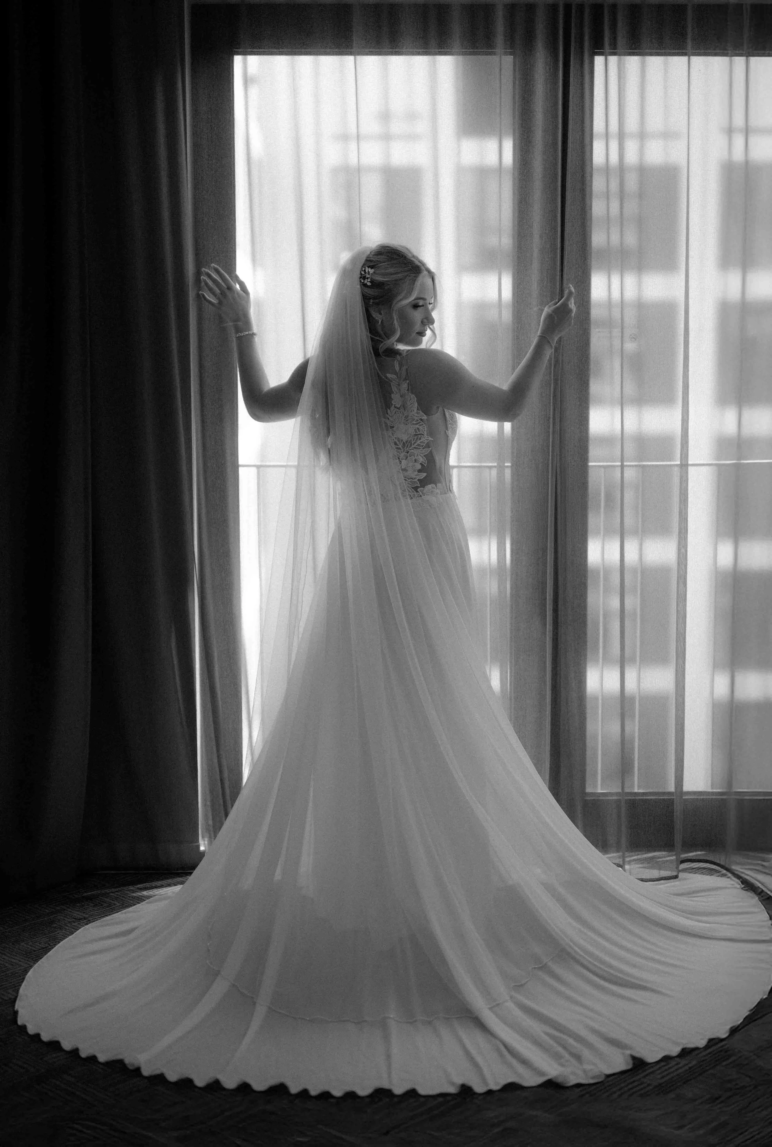 Braut in Hochzeitskleid mit Schleier vor einem großen Fenster mit Vorhängen, schwarz-weiß. Es ist das Getting Ready der Braut im Hotel in Köln. 