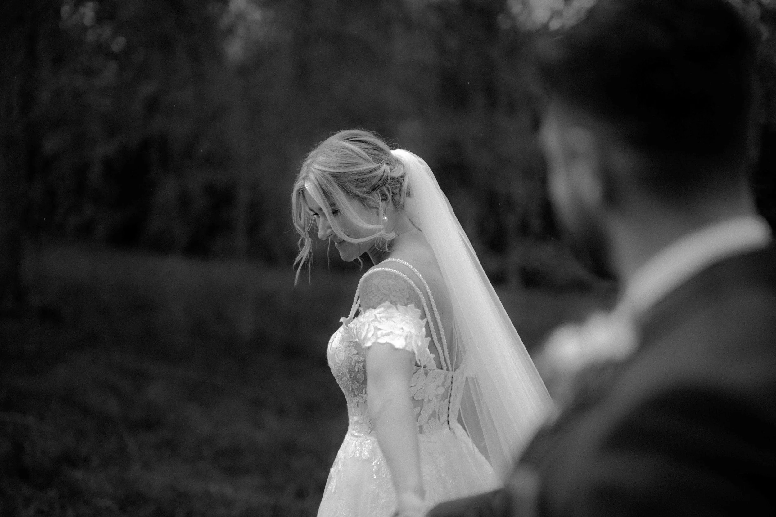 Braut in Hochzeitskleid und Schleier steht im Freien, während sie nach unten schaut, während der Bräutigam im Vordergrund unscharf im Hintergrund steht. Die Hochzeit findet in Düsseldorf, Nähe Köln statt. 