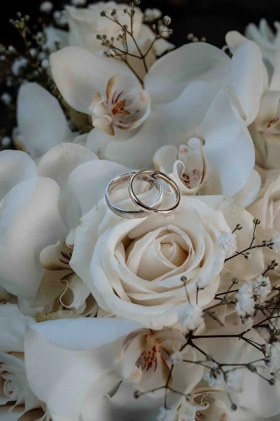 Zwei Eheringe auf einer weißen Rose, umgeben von weißen Orchideen und kleinen Blumen in einer Hochzeitsdeko. Die Hochzeit findet in Düsseldorf, Nähe Köln statt. 