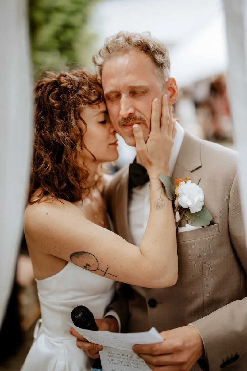 Ein Brautpaar bei einer emotionalen Zeremonie, bei der die Braut den Bräutigam berührt, während er eine Rede hält. Der Bräutigam trägt einen eleganten Anzug mit Blumen im Revers, die Braut trägt ein weißes Kleid mit Tattoos auf dem Arm. Die Hochzeit 