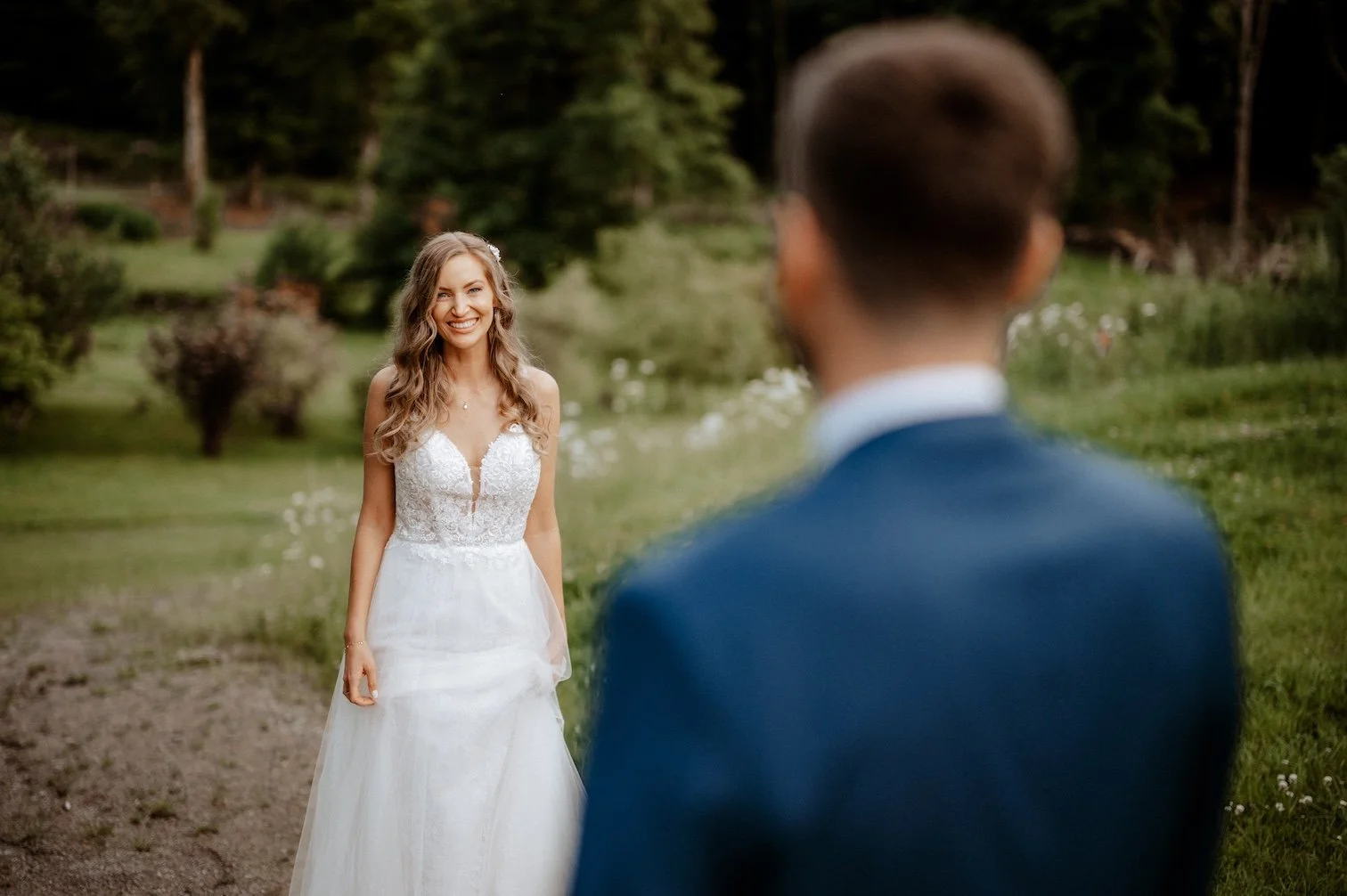 Braut lächelt verliebt und schaut ihren Bräutigam an, der im Vordergrund unscharf ist, in einem grünen Garten bei Sonnenuntergang. Die Hochzeit findet in Düsseldorf, Nähe Köln statt. 