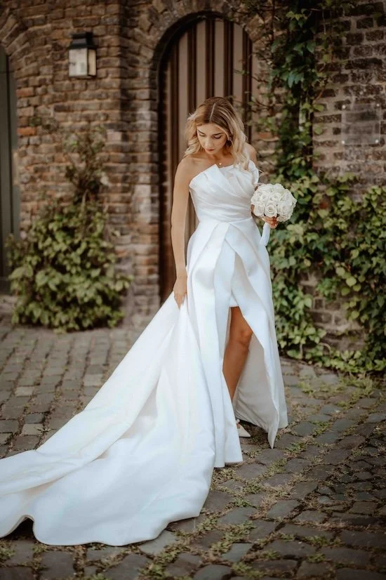 Junge Braut in weißem Hochzeitskleid mit Blumenstrauß vor einer Backsteinmauer und Pflanzen im Hintergrund. Die Hochzeit findet in Düsseldorf, Nähe Köln statt. 