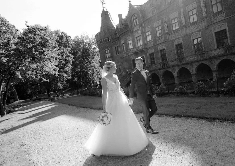 Braut in Hochzeitstracht hält die Hand ihres Bräutigams, während sie einen Blumenstrauß trägt, vor einem alten Schloss bei Sonnenlicht. Die Hochzeit findet in Düsseldorf, Nähe Köln statt. 