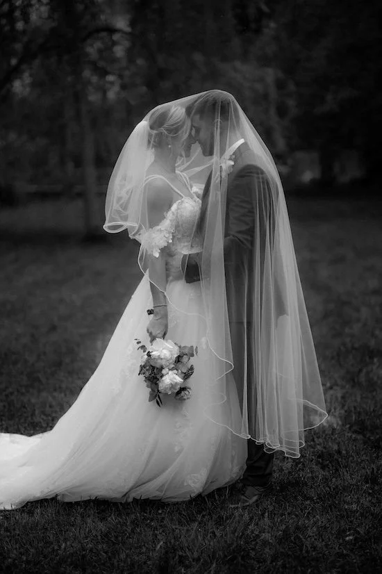 Braut und Bräutigam stehen sich mit Tüllhochzeitskleid und Anzug gegenüber, unter dem Schleier, der sie verbindet, im Freien, mit Brautstrauß in der Hand. Das Brautpaar befindet sich in Bergisch Gladbach bei Köln. 