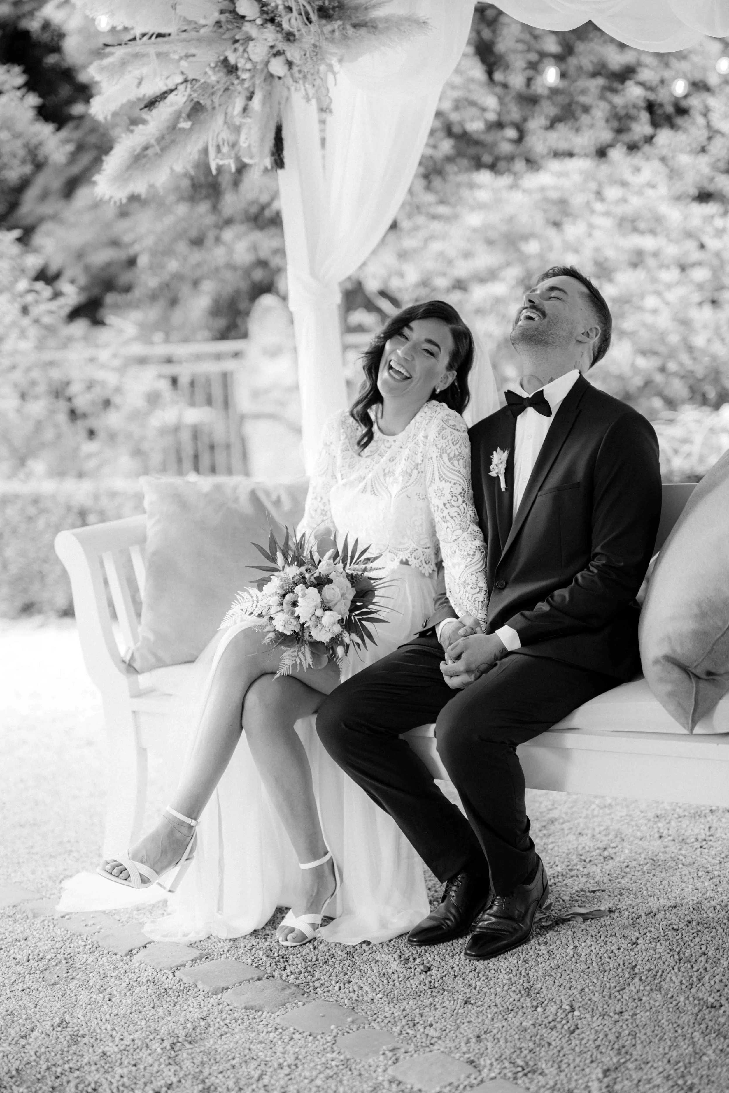Ein lachendes Brautpaar sitzt auf einer Bank bei einer Hochzeit im Freien, die Braut in einem weißen Kleid mit Blumenstrauß, der Bräutigam in einem schwarzen Anzug mit Fliege. Die Hochzeit findet im Schloss Grünewald bei Solingen in der Nähe von Düss