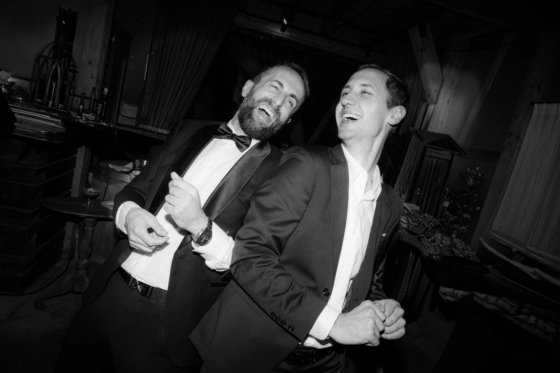 Zwei Männer in eleganter Kleidung, lachen und haben Spaß auf einer Party in einem Chalet, schwarz-weiß Foto von einer Hochzeit. Der Ort ist in Düsseldorf bei einer Hochzeit. 