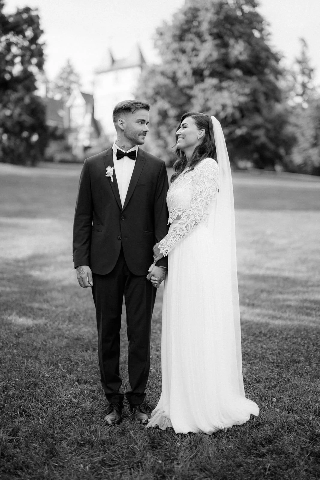 Ein Brautpaar steht sich gegenüber auf einer Wiese, hält Händchen und blickt sich verliebt an, im Hintergrund Bäume und ein Gebäude. Die Hochzeit findet im Schloss Grünewald in Solingen, bei Düsseldorf, Nähe Köln statt. 