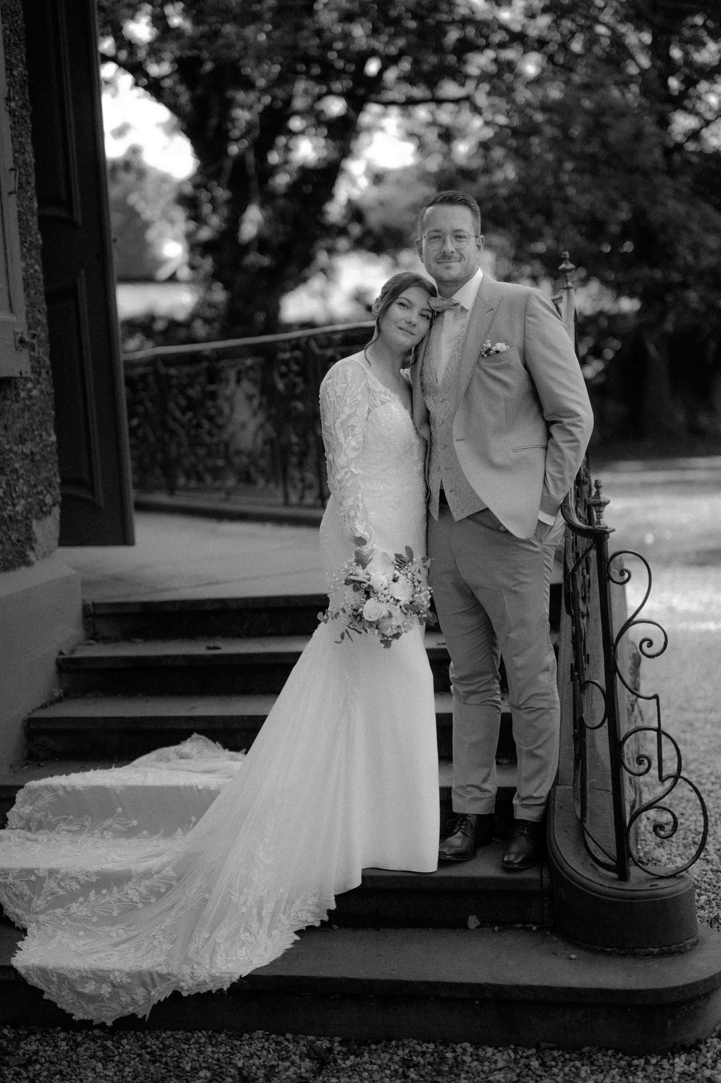 Ein Brautpaar steht eng zusammen auf einer Treppe im Freien, die Frau trägt ein Hochzeitskleid mit langem Schleier und hält einen Blumenstrauß, der Mann trägt einen Anzug. Im Hintergrund sind Bäume und eine Zaun. Das Bild ist in Schwarzweiß. Die Hoch