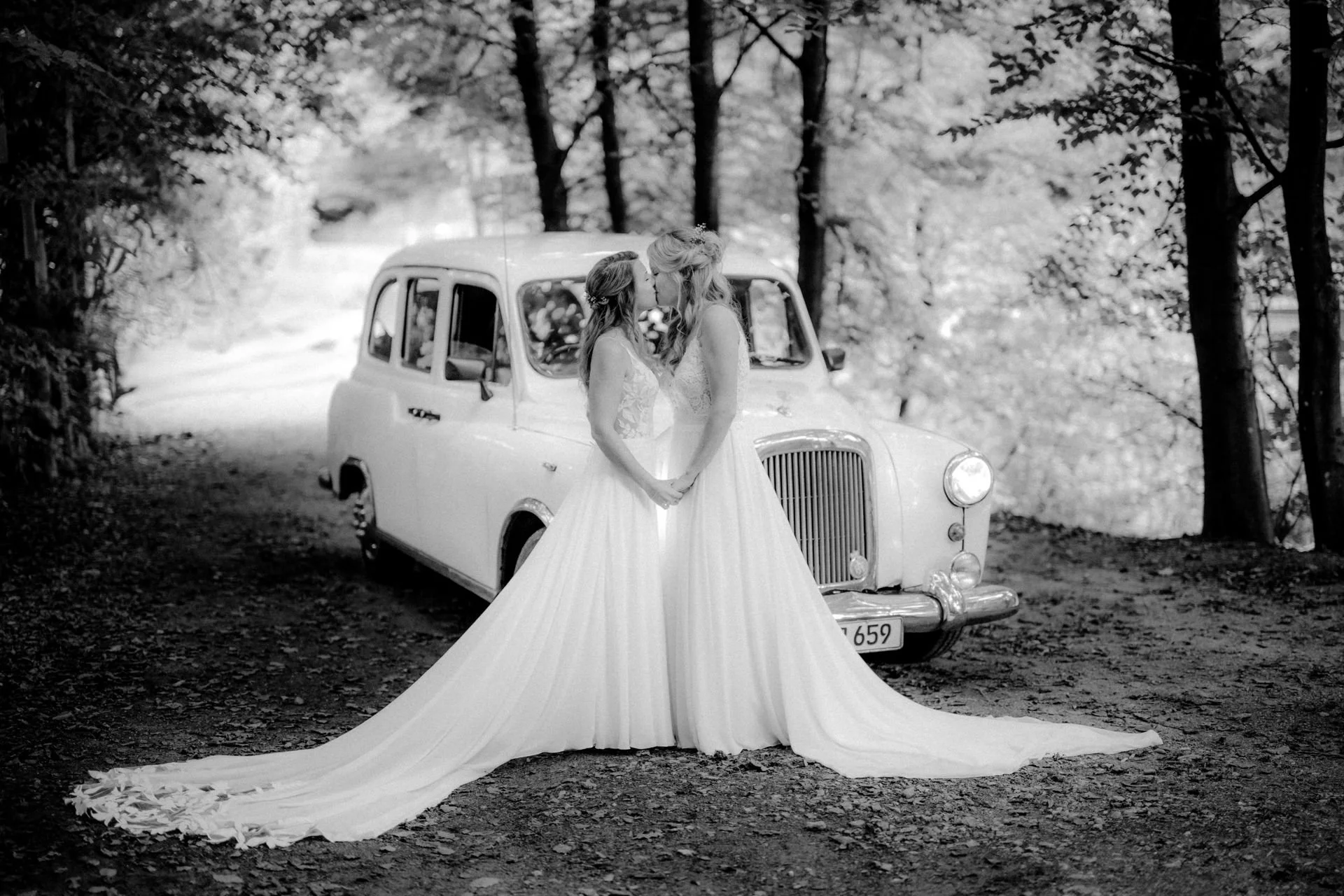 Zwei Bräute in Hochzeitskleidern küssen sich vor einem alten weißen Auto in einem Wald. Die Hochzeit findet in Leichlingen im Sinneswald bei Düsseldorf, Nähe Köln statt. 