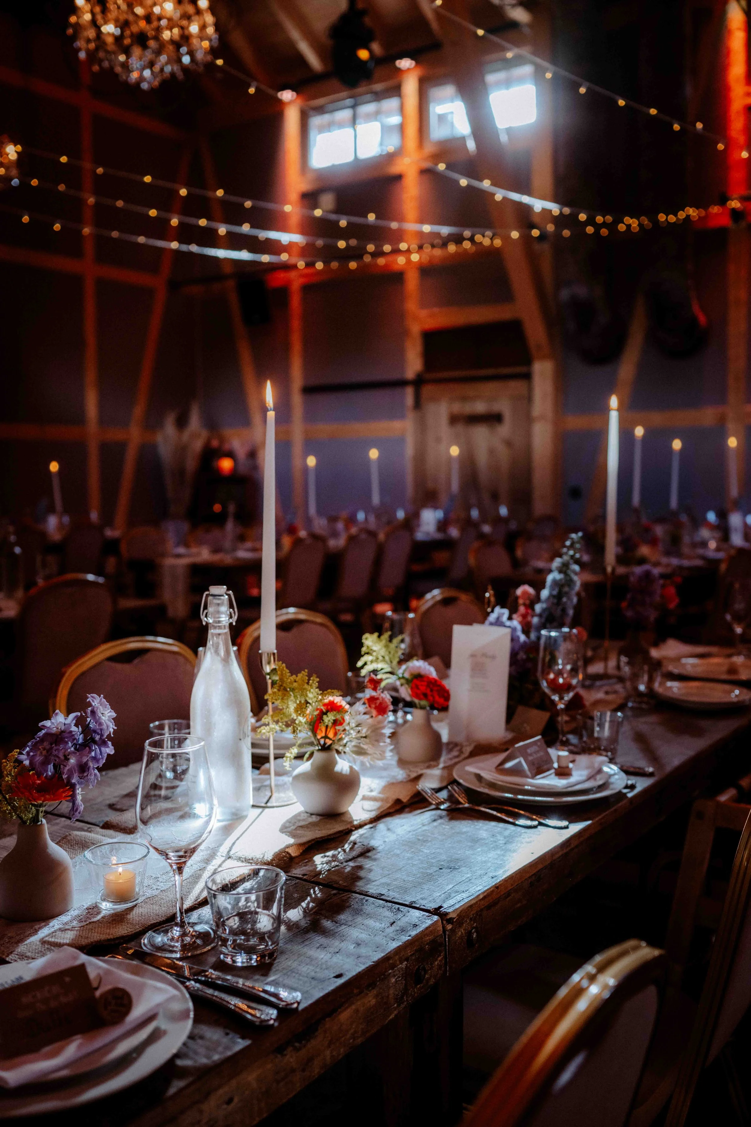 Elegante festliche Tafel mit Kerzen, Blumen und Gläsern in einem rustikalen Raum mit Holzdecken und Lichterketten.Die Hochzeit findet in Düsseldorf, Nähe Köln statt. 