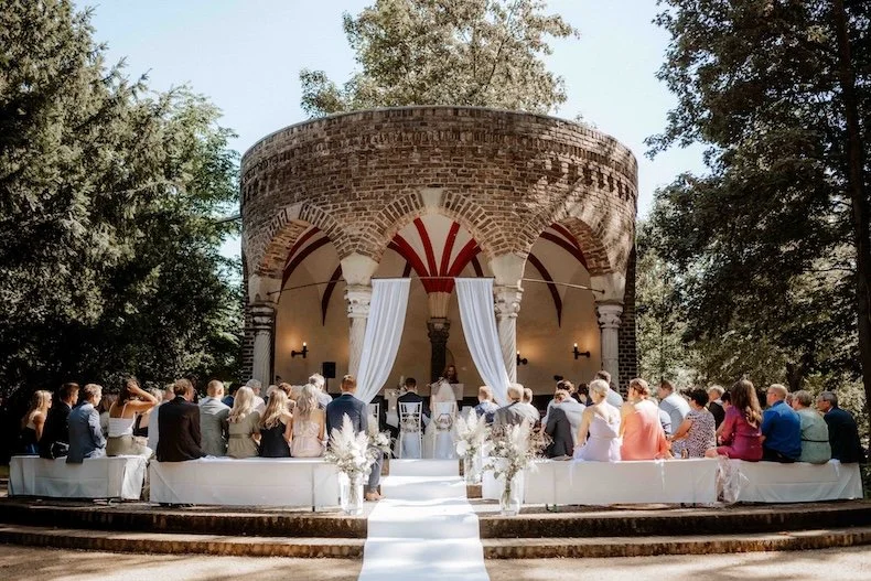 Eine Hochzeitszeremonie im Freien vor einer historischen Steinarche, umgeben von Bäumen, mit Gästen in formeller Kleidung, einer Braut in weißem Kleid und einem Bräutigam, die an einem Altar stehen. Die Hochzeit findet in Düsseldorf, Nähe Köln statt.