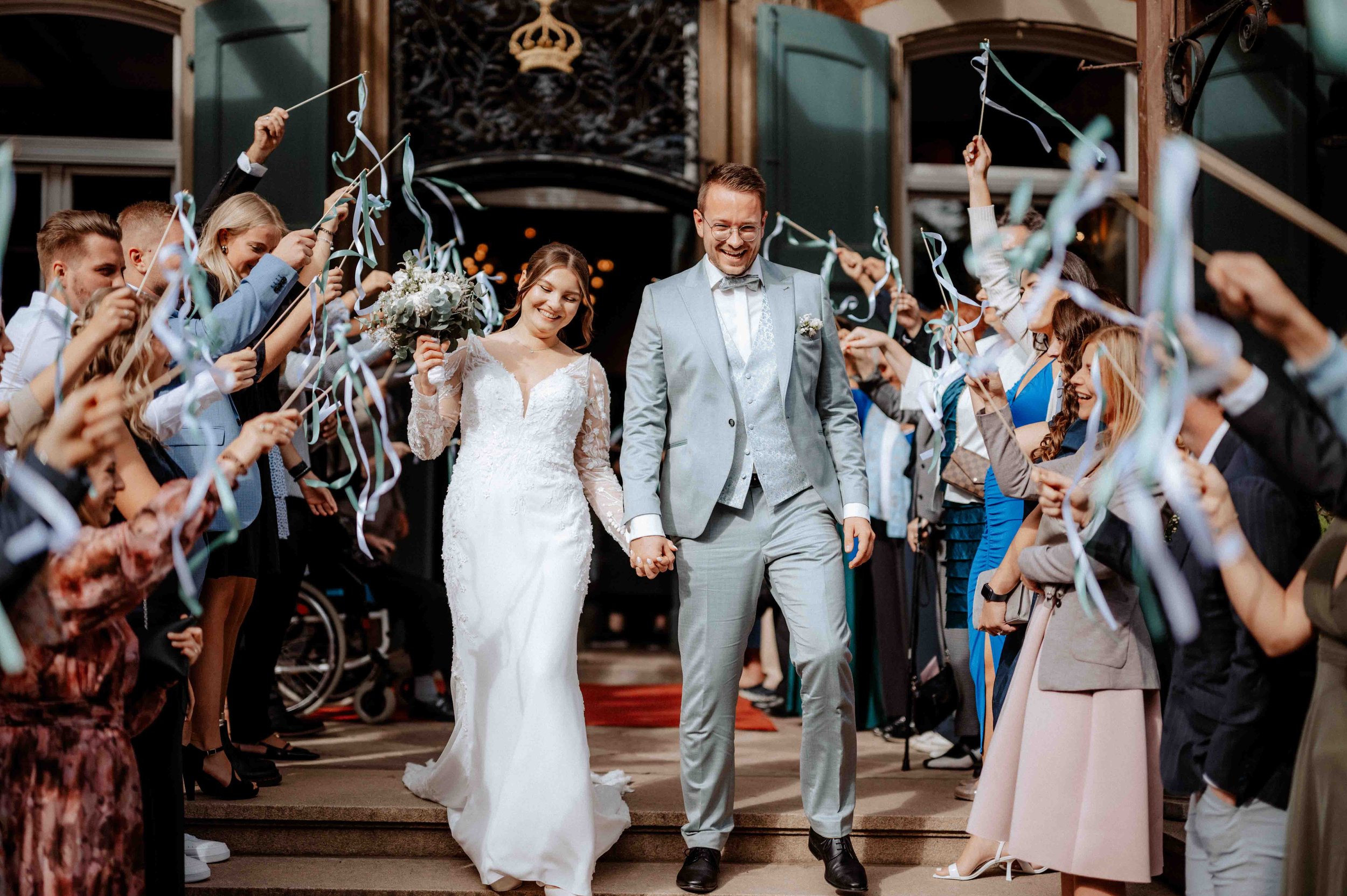 Ein glückliches Brautpaar in stilvoller Hochzeitskleidung, umgeben von Freunden und Familie, die ihnen mit Papierbändern zujubeln und feiern, vor einer alten, grünen Tür. Die Hochzeit findet im Schloss Eicherhof in Leichlingen, Nähe Köln statt. 