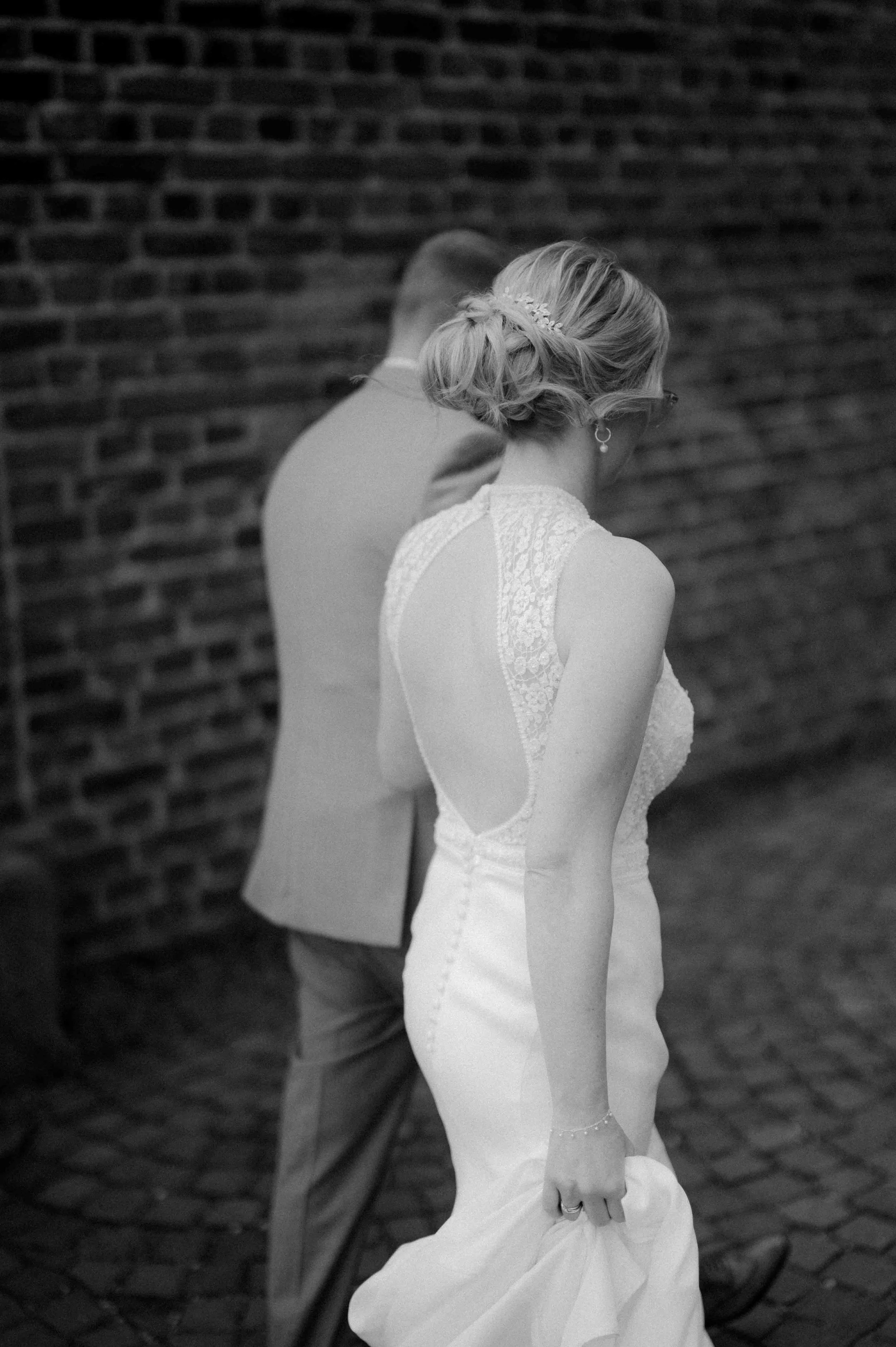 Braut in Hochzeitskleid mit Hochsteck-Frisur, neben Brautführer in Anzug, an einer Backsteinwand. Die Hochzeit findet in Düsseldorf beim Schloss Benrath statt. 