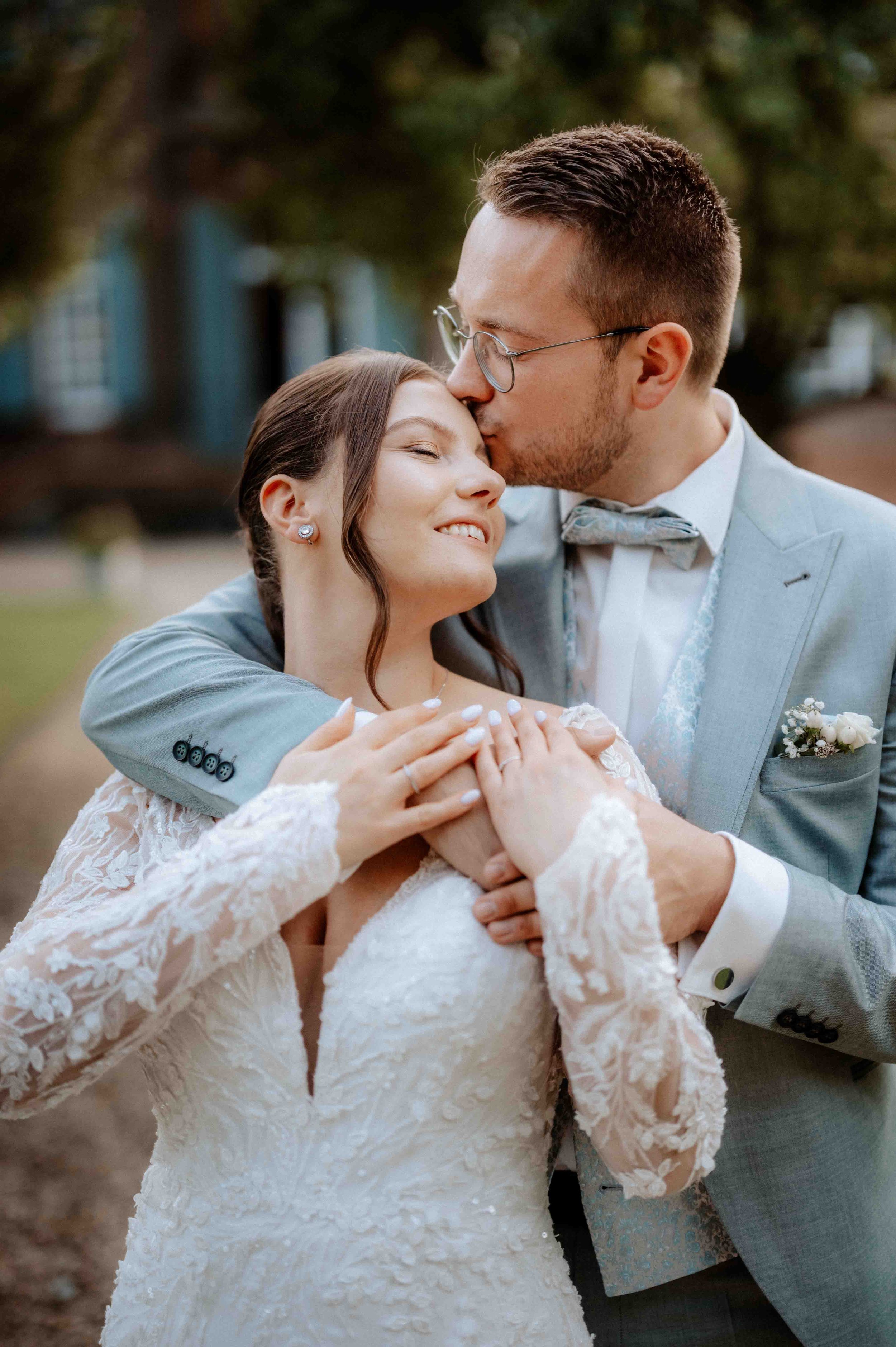 Ein Brautpaar umarmt sich im Freien, der Bräutigam küsst die Braut auf die Stirn, beide sind in eleganter Hochzeitskleidung, die Braut trägt ein Spitzenkleid, der Mann einen Anzug mit Fliege. Die Hochzeit findet im Schloss Eicherhof in Leichlingen be