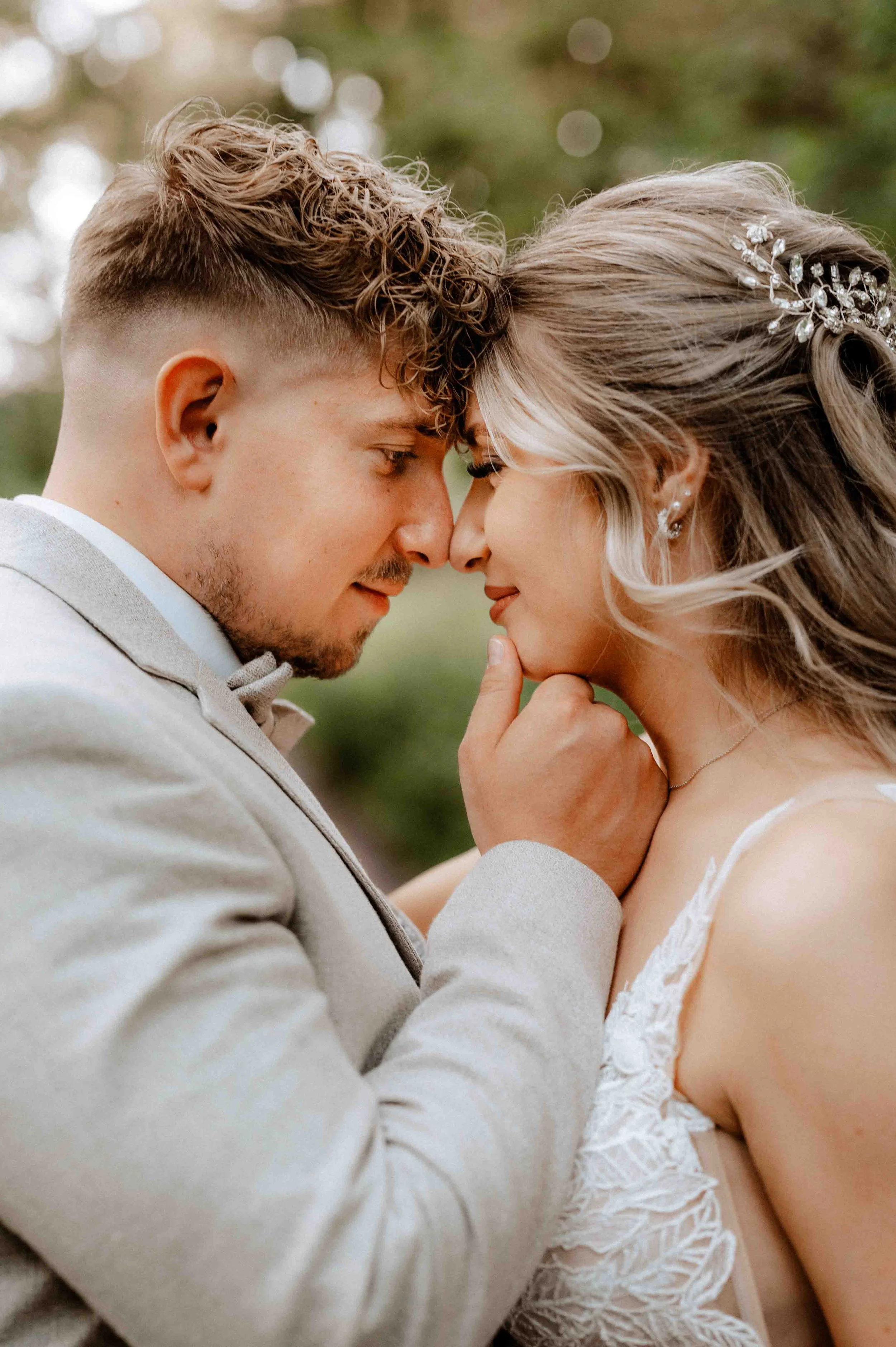 Ein heiratendes Paar steht sich nahe und berührt sich mit den Stirn. Die Frau trägt ein makelloses Brautkleid mit Spitze und hat lange, gewellte Haare mit Haarschmuck. Der Mann trägt einen hellen Anzug. Sie scheinen einen Moment voller Liebe und Verb