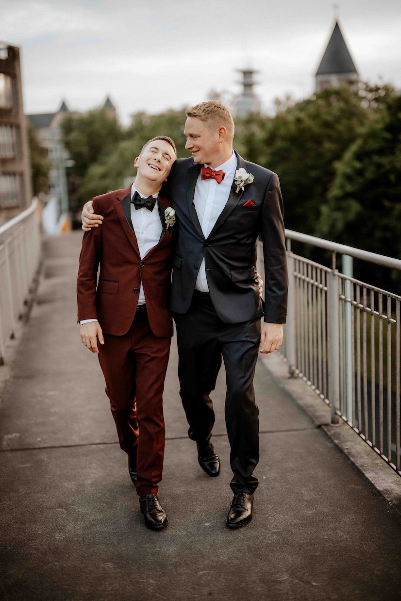 Zwei Männer in Anzügen, die sich auf einer Brücke umarmen, während sie lächeln. Einer trägt einen dunkelroten Anzug, der andere einen schwarzen Anzug, beide mit Fliegen. Im Hintergrund sind Bäume und ein Gebäude mit eine Turm sichtbar. Die Hochzeit f