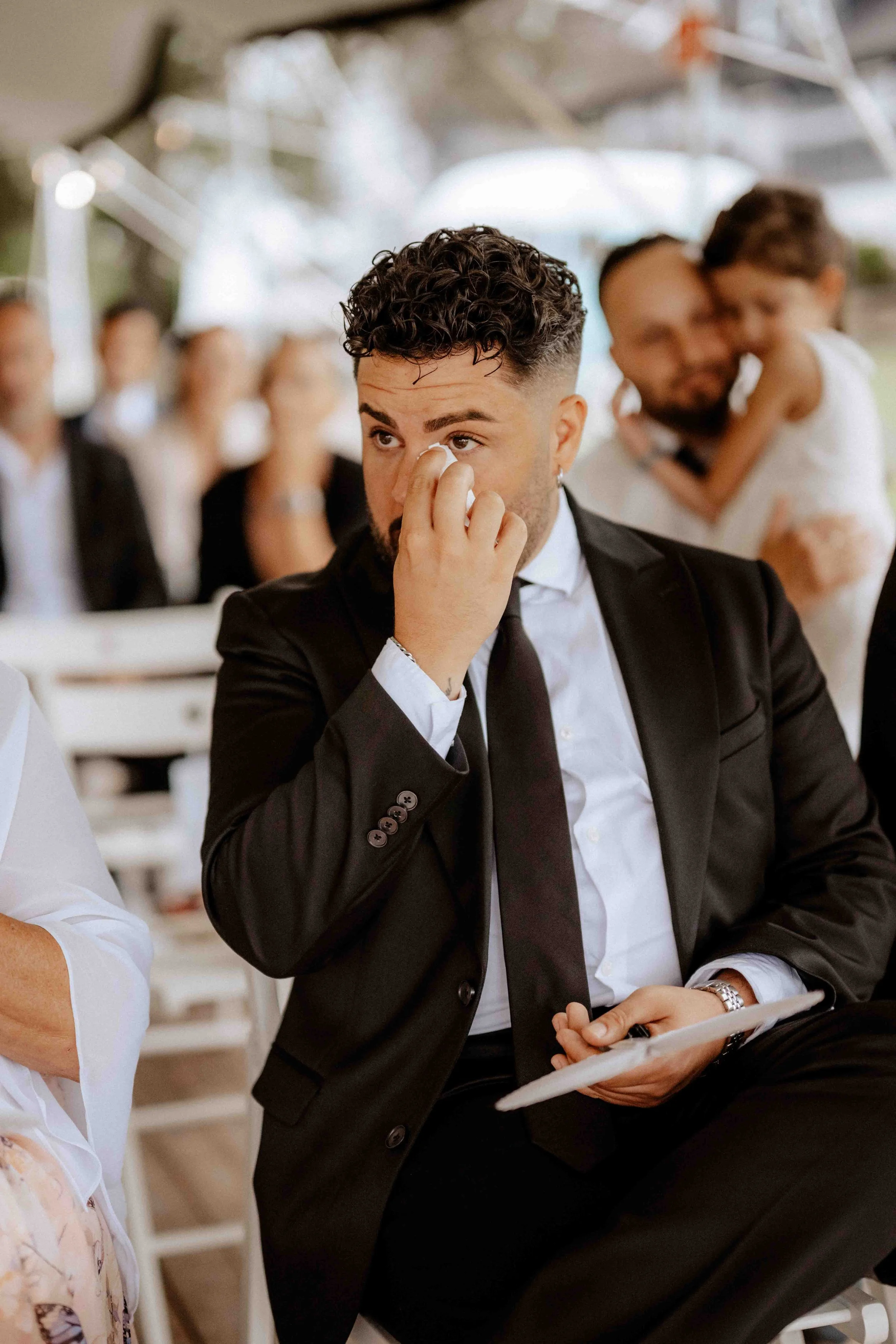 Ein junger Mann in einem schwarzen Anzug, der bei einer Veranstaltung emotional ist und die Tränen abwischt, während andere Menschen im Hintergrund lächeln und sich umarmen. Die Hochzeit findet in Düsseldorf, Nähe Köln statt. 