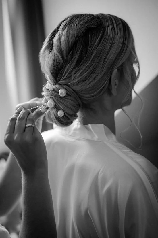 Frau mit elegant hochgestecktem Haar, das mit Perlen verziert ist, während sie sich an einem Spiegel schminkt. Die Hochzeit findet in Düsseldorf, Nähe Köln statt. 