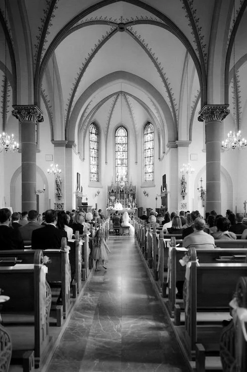 Schwarz-weiß-Fotografie einer Kirche während einer Trauung, mit Gästen, Bänken und einem Altar im Hintergrund. Die Hochzeit findet in Langenfeld bei Düsseldorf, Nähe Köln statt. 