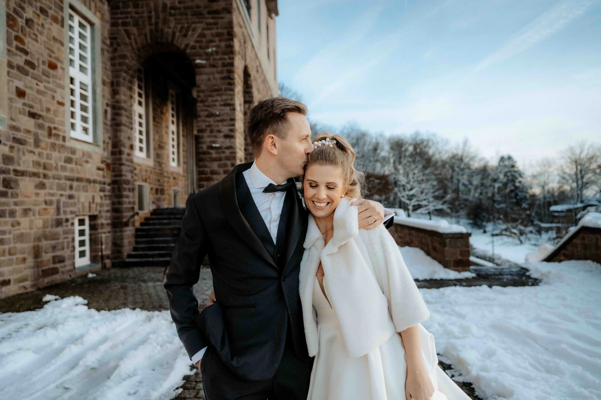 Ein lächelndes Brautpaar in formeller Kleidung im Winter vor einem Backsteingebäude, umgeben von Schnee, beim Fotoshooting. Die Hochzeit findet in Bergisch Gladbach im Kardinal Schulte Haus in der Nähe von Köln statt. 