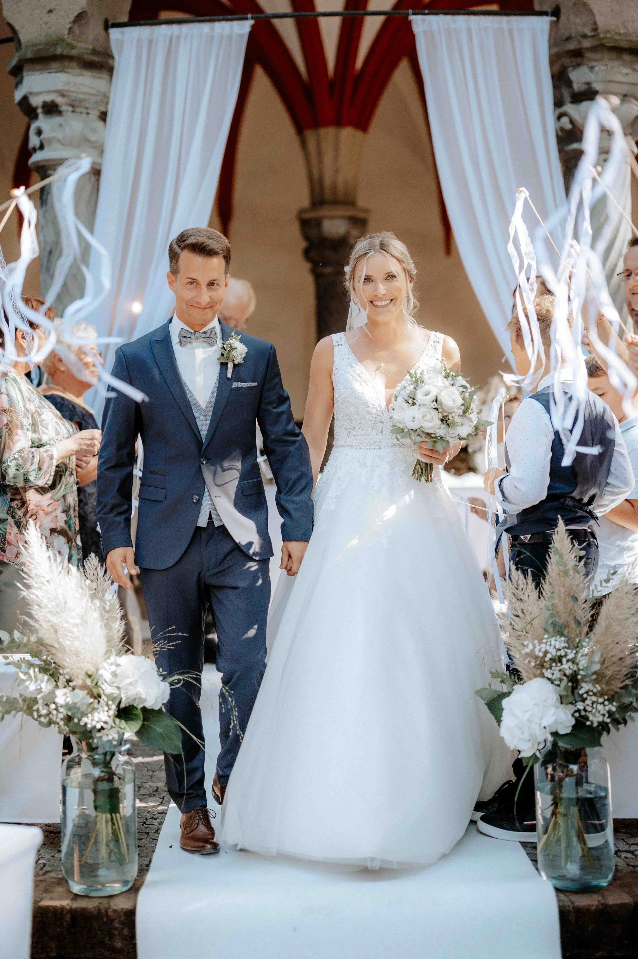 Ein Brautpaar bei der Hochzeit, die Braut trägt ein weißes Brautkleid und hält einen Blumenstrauß, der Bräutigam trägt einen dunkelblauen Anzug mit Fliege. Sie gehen Hand in Hand, umgeben von Blumen und Gästen, im Hintergrund eine architektonische Ku