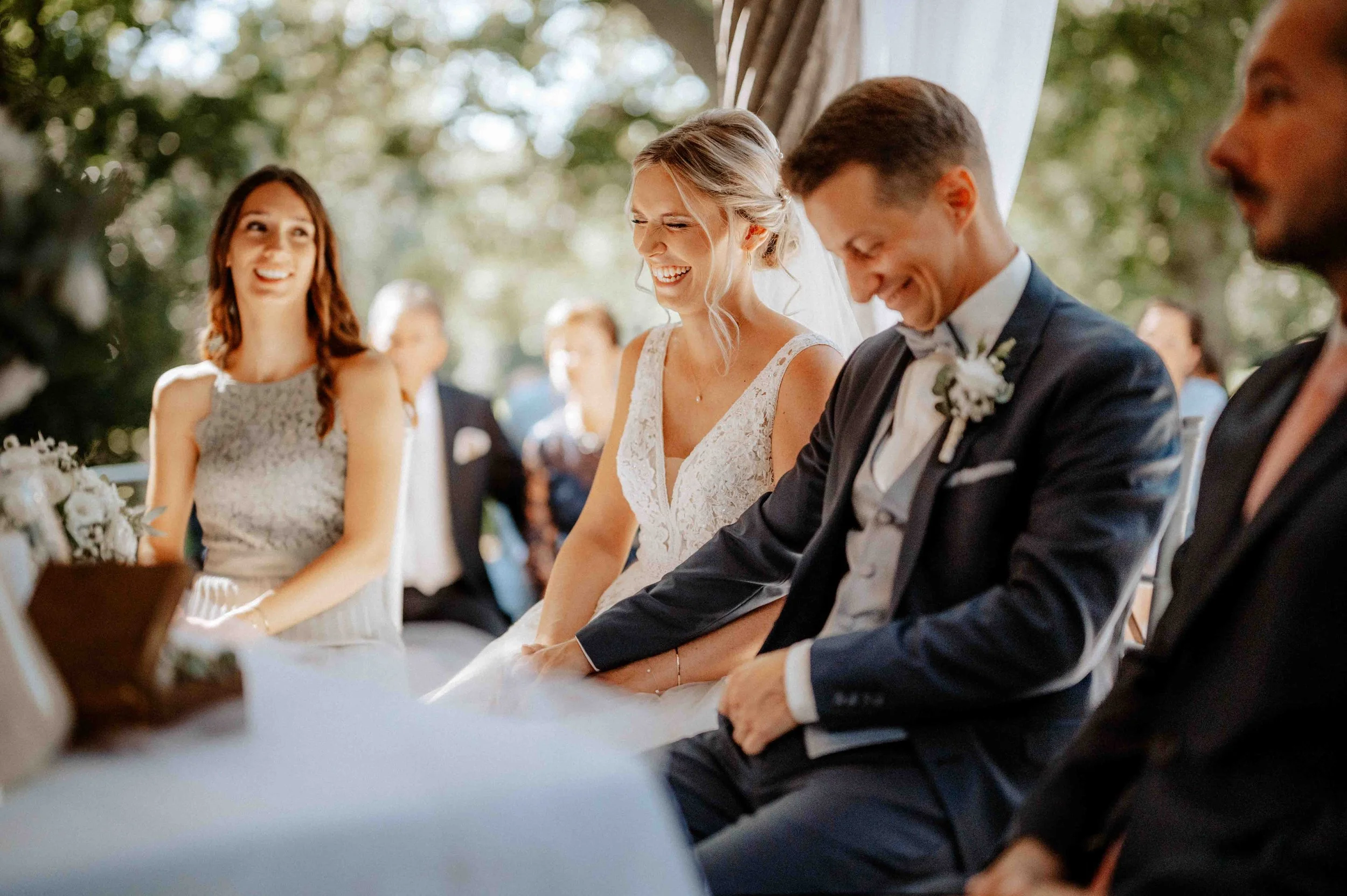 Hochzeitspaar sitzt lachend bei der Trauung, umgeben von Gästen im Freien, Hintergrund aus Bäumen. Die Hochzeit findet im Schloss Paffendorf in Bergheim Nähe Köln statt. 